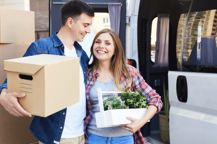 assurance-menage-et-assurance-responsabilite-civile-comment-faire-des-economies Couple souriant en plein déménagement, portant un carton et des plantes.